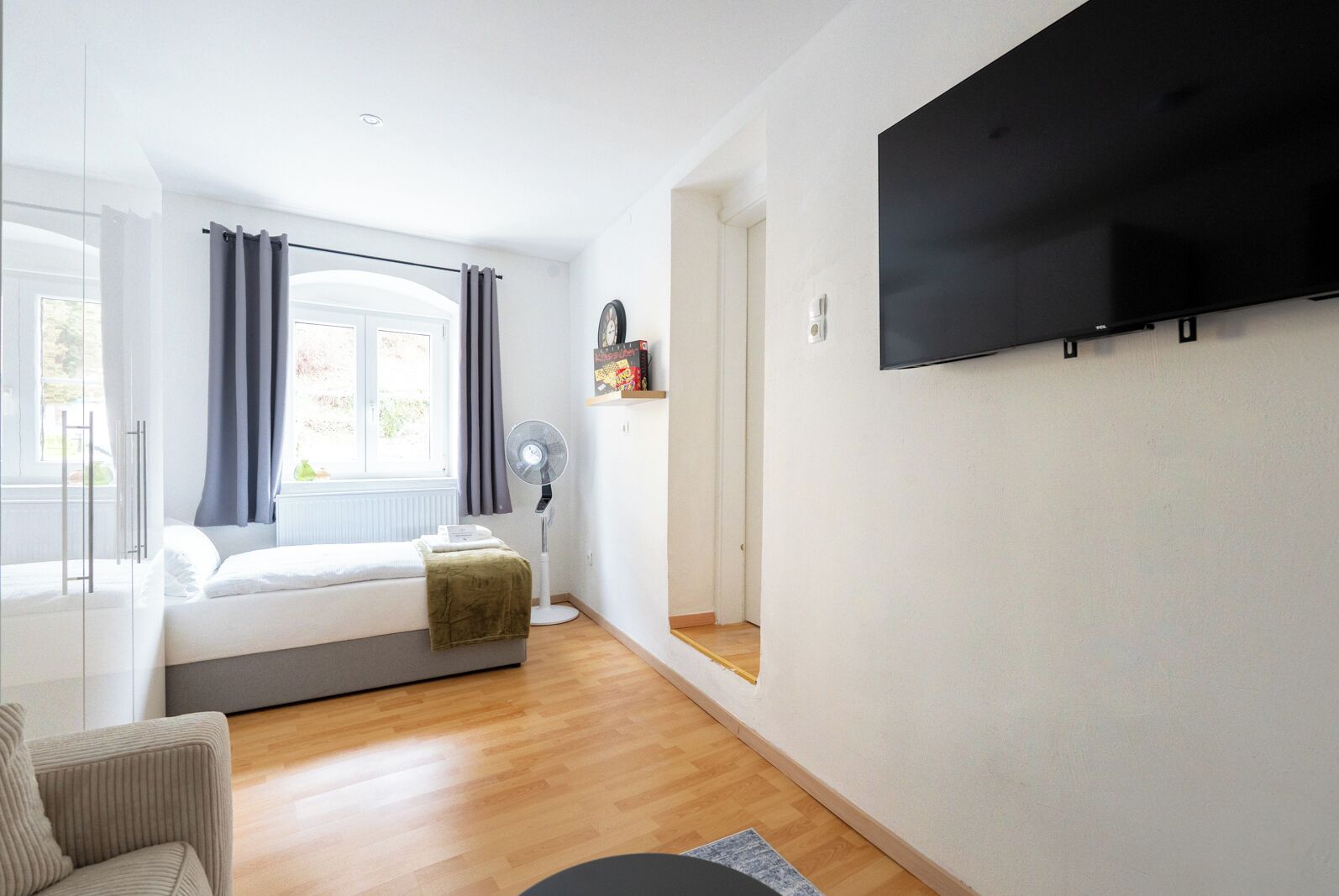 Un dormitorio moderno con cama, ventilador de pie, televisión montada en la pared y ventana con cortinas.