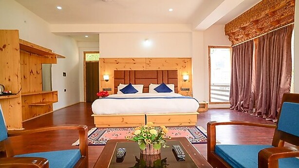 A hotel room with a large bed, wooden headboard, two bedside lamps, a wooden desk, and a large window with curtains. - Hotel Paro Residency (Leh)