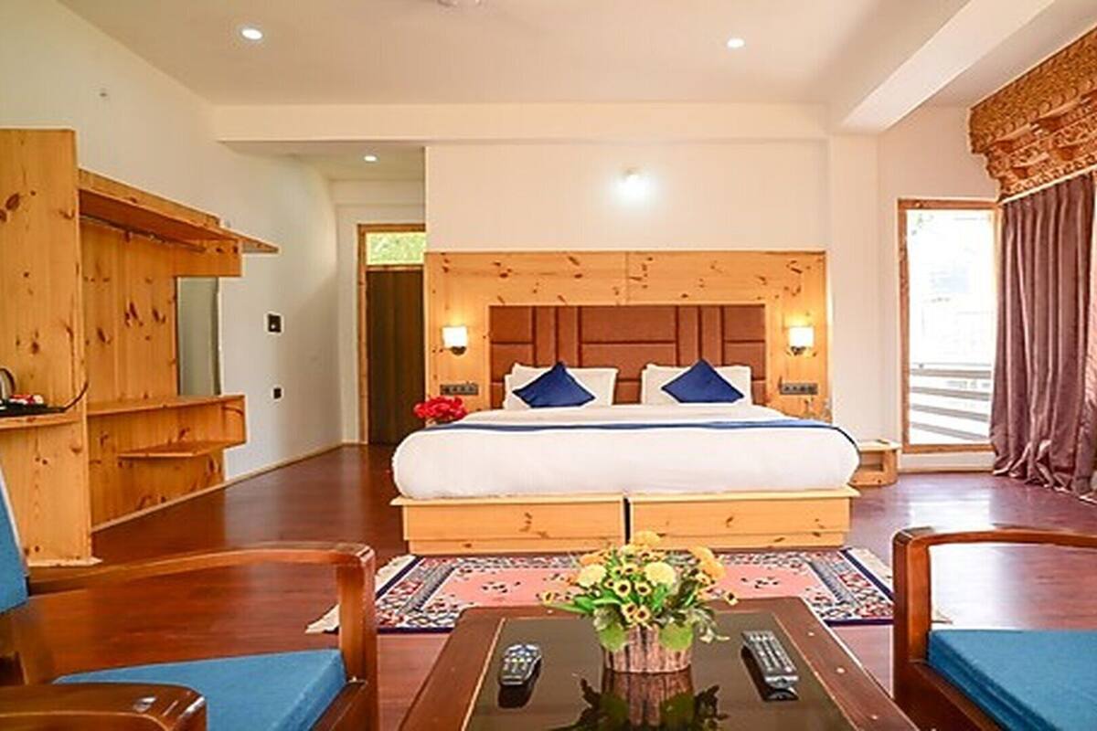A hotel room with a large bed, wooden headboard, two bedside lamps, a wooden desk, and a large window with curtains.