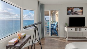 Habitación de hotel con un ventanal grande con vistas al mar, un telescopio, un escritorio con libros, un televisor y un balcón con sillas azules.