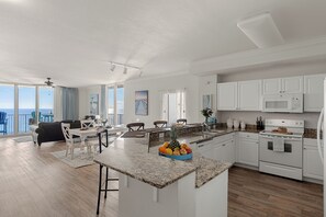A modern kitchen with white cabinets, a granite countertop, and a central island with fruit. - Beachfront Condo + Resort Pool + Spa Amenities + Movie Theater + Game Room (Panama City Beach)