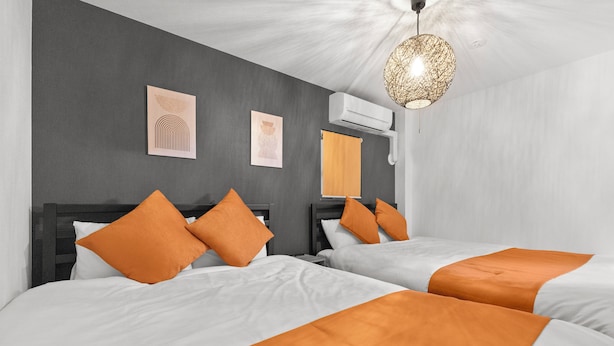 Ein Hotelzimmer mit zwei Betten, jeweils mit weißer Bettwäsche und orangefarbenen Akzenten. An den Wänden sind Klimaanlagen angebracht und eine Deckenleuchte ist vorhanden.