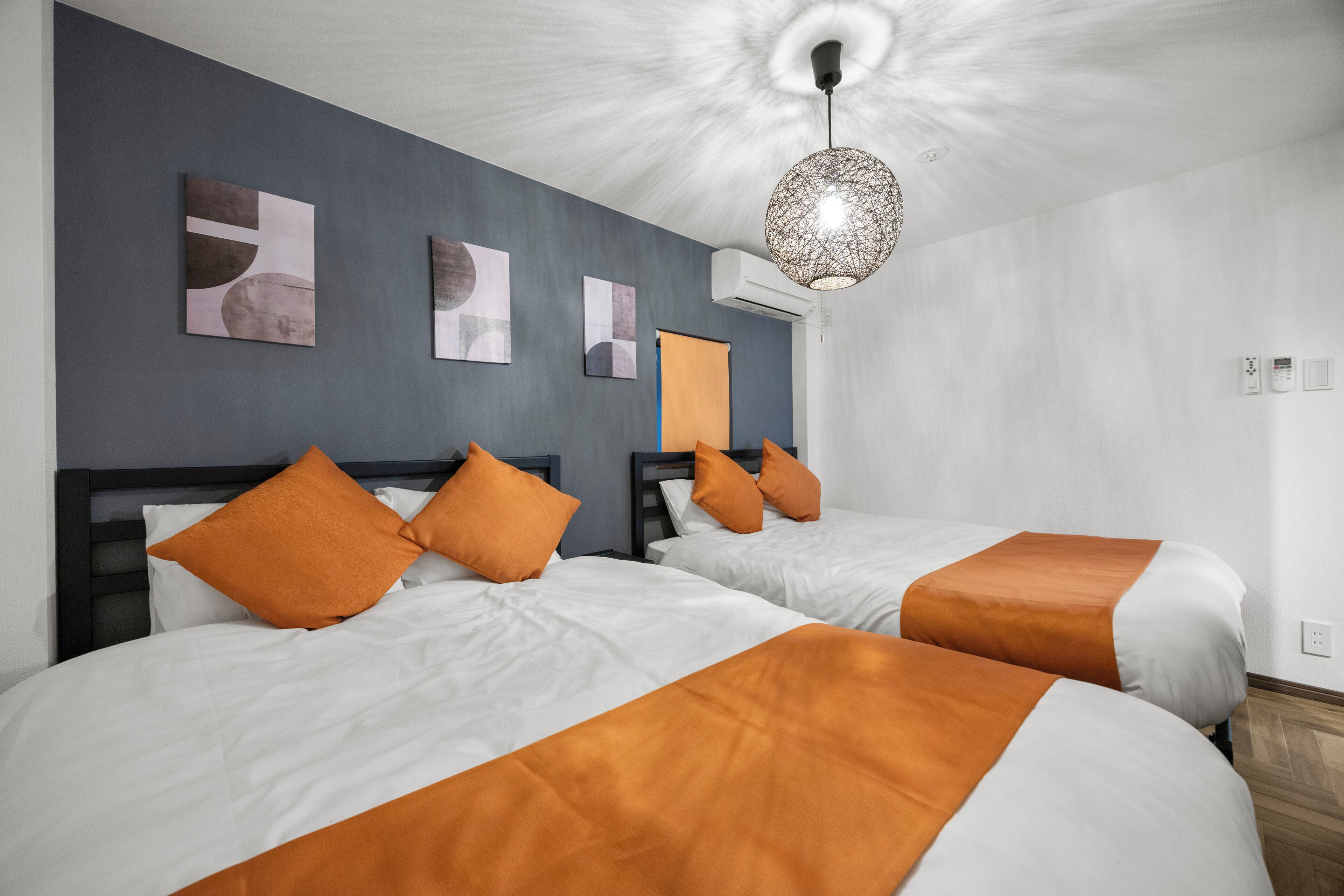 A hotel room with two beds, each with white bedding and orange accents, a modern pendant light, and artwork on the walls.