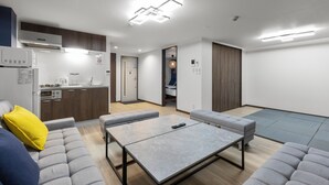 Ein modernes, kompaktes Wohnzimmer mit Küchenzeile, grauen Sofas, einem kleinen Esstisch und einer Schiebetür, die zum Schlafzimmer führt.