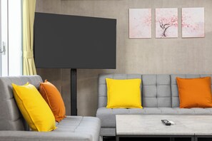 A modern living room with a grey sofa, yellow and orange cushions, a flat-screen TV, and a coffee table.