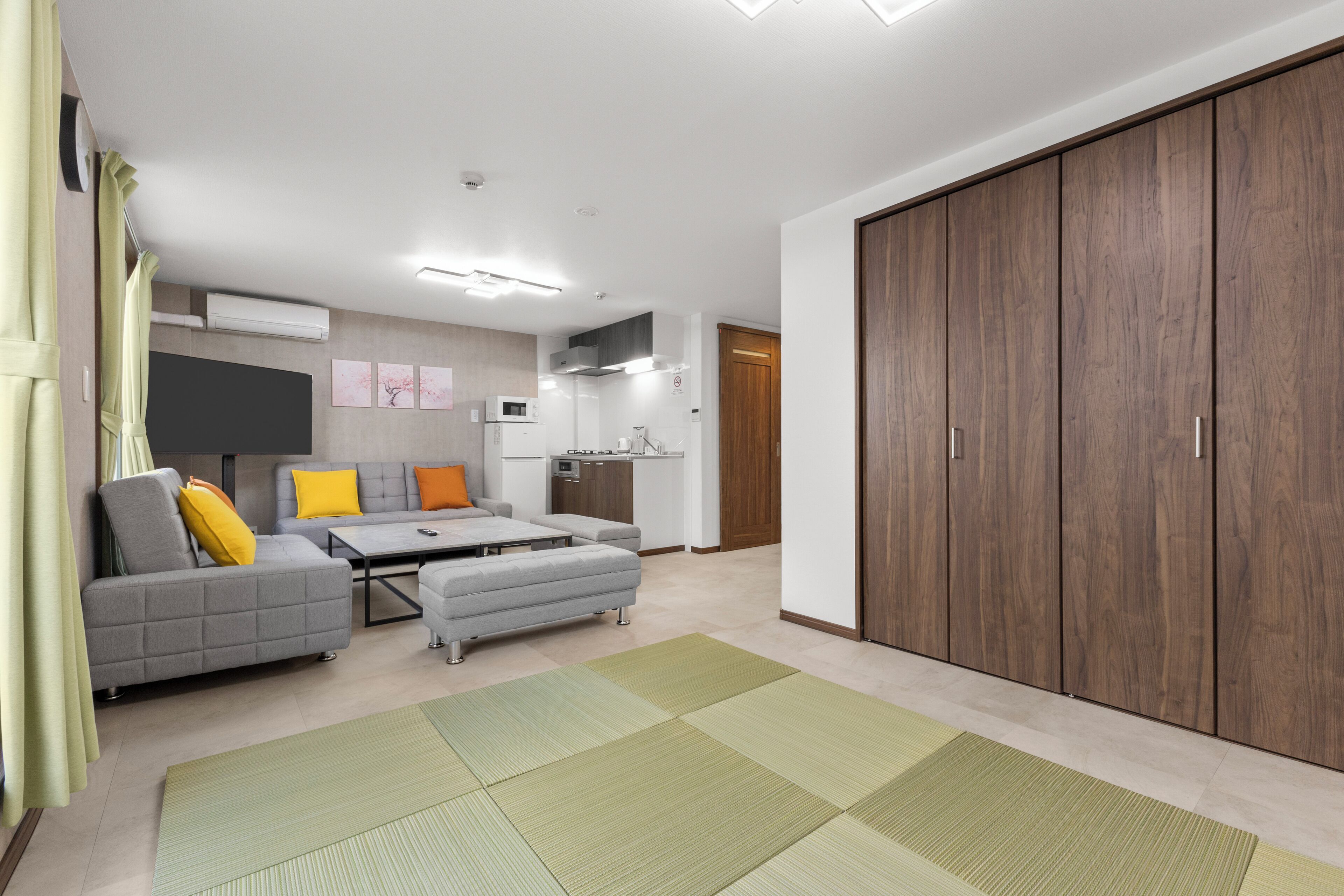 Een moderne woonkamer met een bank, voetenbankje en een eettafel. Er is een keukenhoek met een magnetron en kastjes.