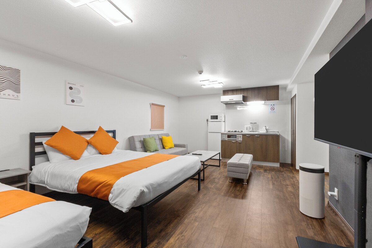 A modern hotel room with two beds, a kitchenette, and a TV.