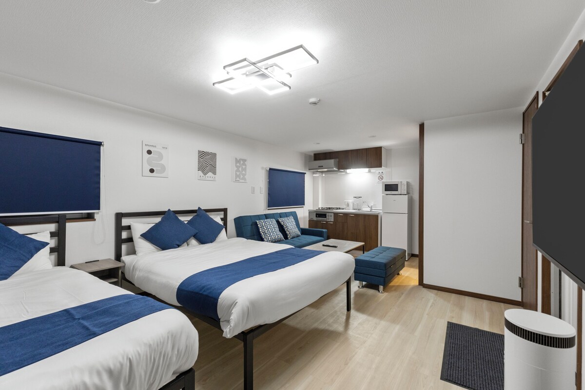 A modern hotel room with two beds, a sofa, a small kitchenette, and a flat-screen TV.