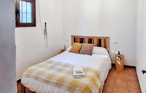 Un dormitorio con cabecera de madera, colcha a cuadros, dos almohadas, una mesita de noche con lámpara y una ventana con barrotes.