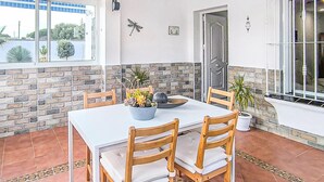 Un comedor con una mesa blanca, sillas de madera y un cuenco de flores. Se aprecia una pared de ladrillo y una puerta al fondo.