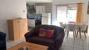 Ett kompakt bostadsutrymme med ett litet kök, en soffa, ett matbord med stolar och en utsikt genom ett fönster med gardiner.