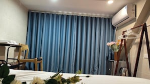 A hotel room with a bed, a chair, a table with a towel, a blue curtain, an air conditioner, a hanger, and a flower vase. - Omeli Homestay (Hanoi)