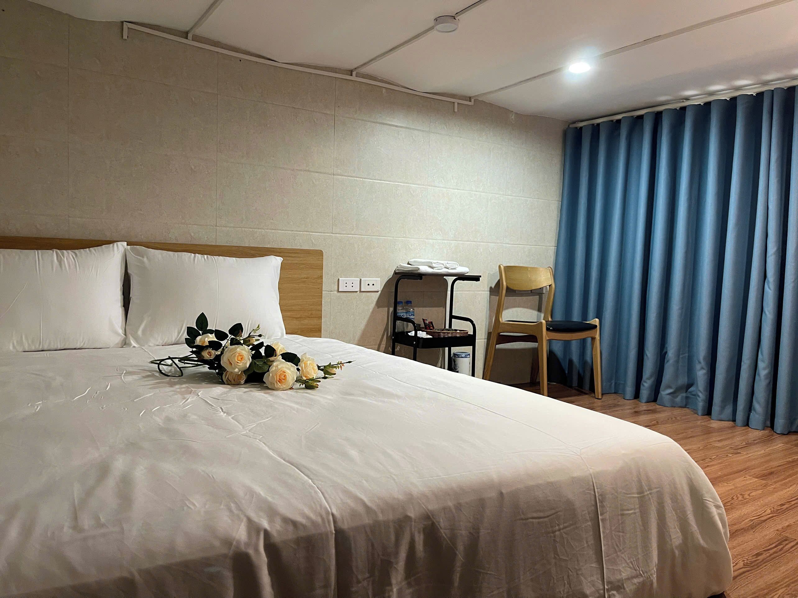 A hotel room with a large bed, a wooden headboard, a desk with a chair, a blue curtain, and a vase of flowers.