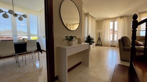 Un vaste espace de vie agrémenté d’une grande fenêtre, d’un miroir rond, d’une console blanche ornée d’objets décoratifs, d’un lampadaire sur trépied et d’un canapé.