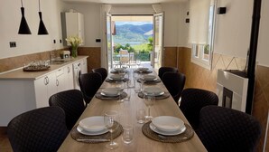 On aperçoit une salle à manger avec une longue table dressée pour six personnes, équipée d’assiettes, de verres et de couverts. Une cuisine, avec un évier et des placards, est visible sur la gauche.
