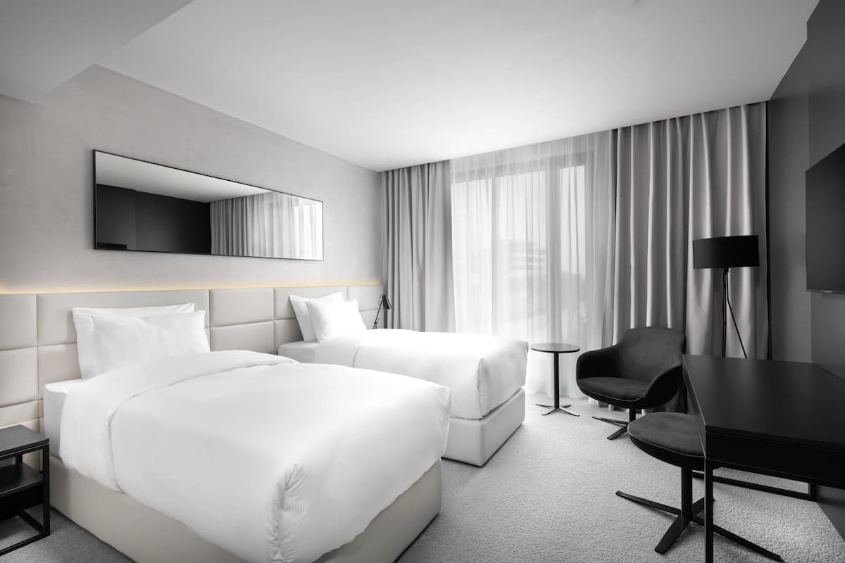 A hotel room with two beds, a desk, a chair, a TV, and a large window with curtains.