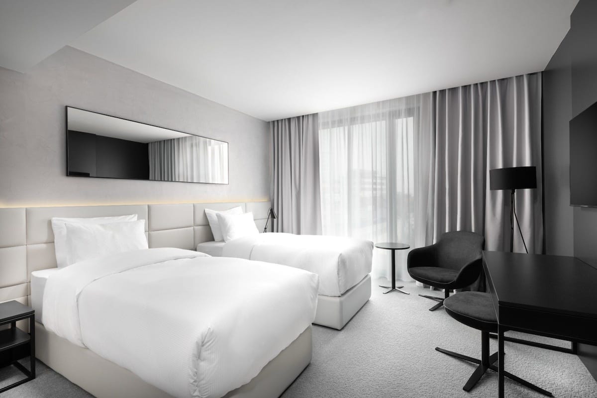 A hotel room with two beds, a desk, a chair, a TV, and a large window with curtains.