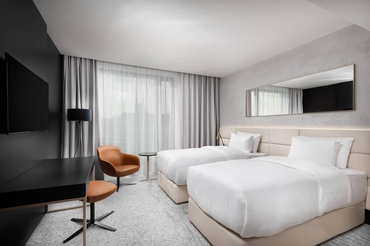 A hotel room with a bed, a desk, a chair, a TV, and a large window with curtains.