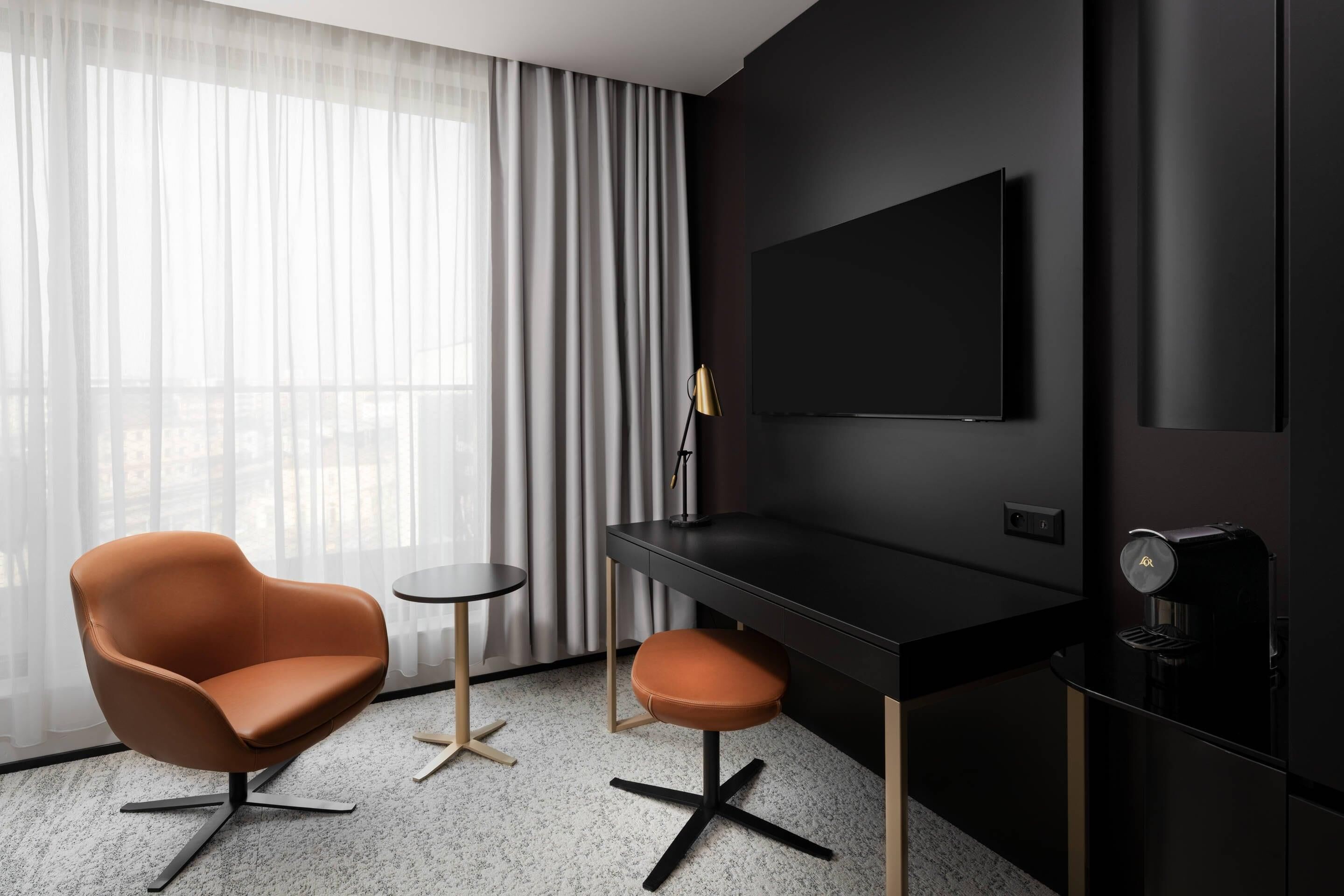 A modern hotel room with a black desk, a leather chair, a small round table, and a large window with sheer curtains.