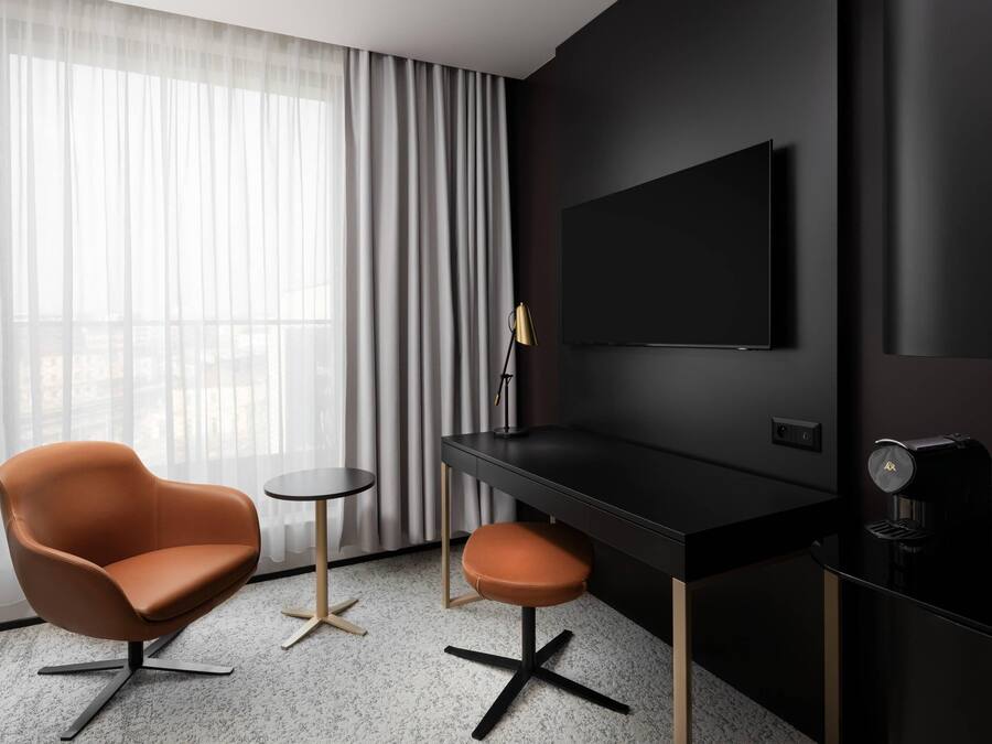 A modern hotel room with a black desk, a leather chair, a small round table, and a large window with sheer curtains.