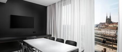 Una sala de conferencias moderna con una gran mesa, sillas negras, un televisor de pantalla plana y vistas a un paisaje urbano a través de amplios ventanales.