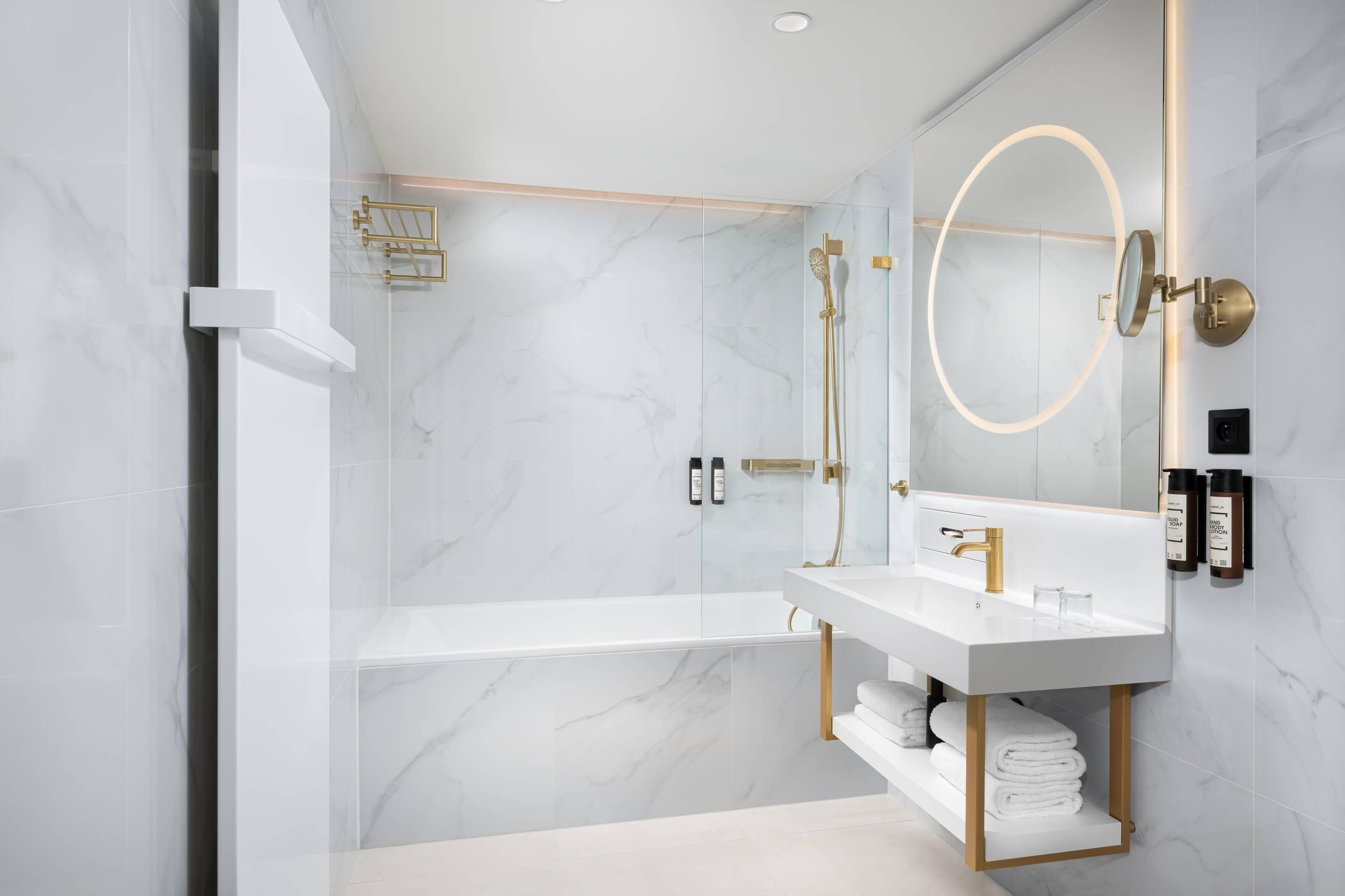 A modern bathroom with a white marble floor, a freestanding bathtub, a vanity with a gold faucet, and a large mirror with a gold frame.