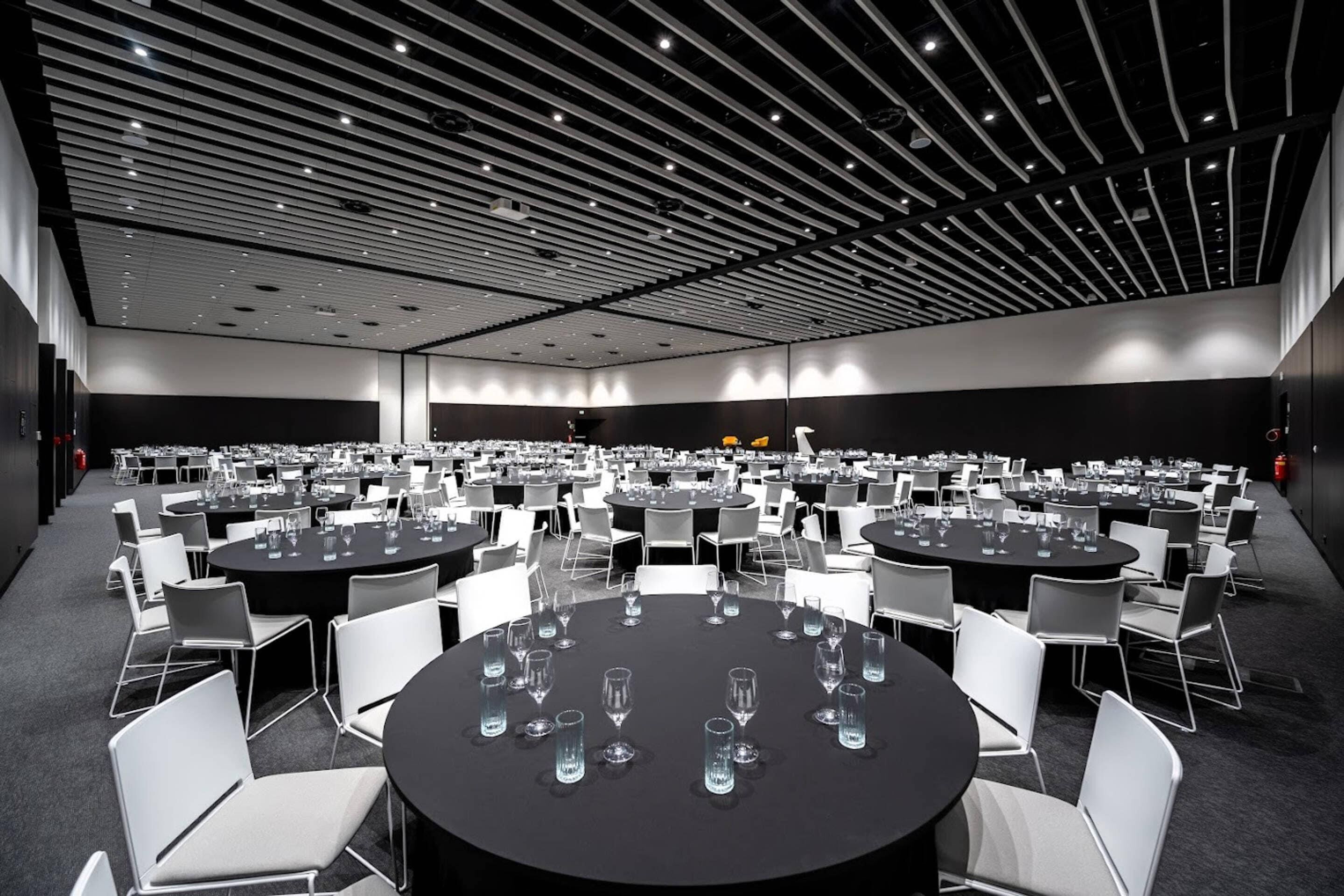 Un amplio salón de conferencias con varias mesas redondas y sillas, techo alto con iluminación lineal y una paleta de colores neutros.
