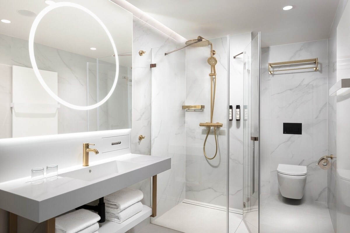 Un baño moderno con ducha envuelta en vidrio, tocador blanco con detalles dorados y un espejo grande.
