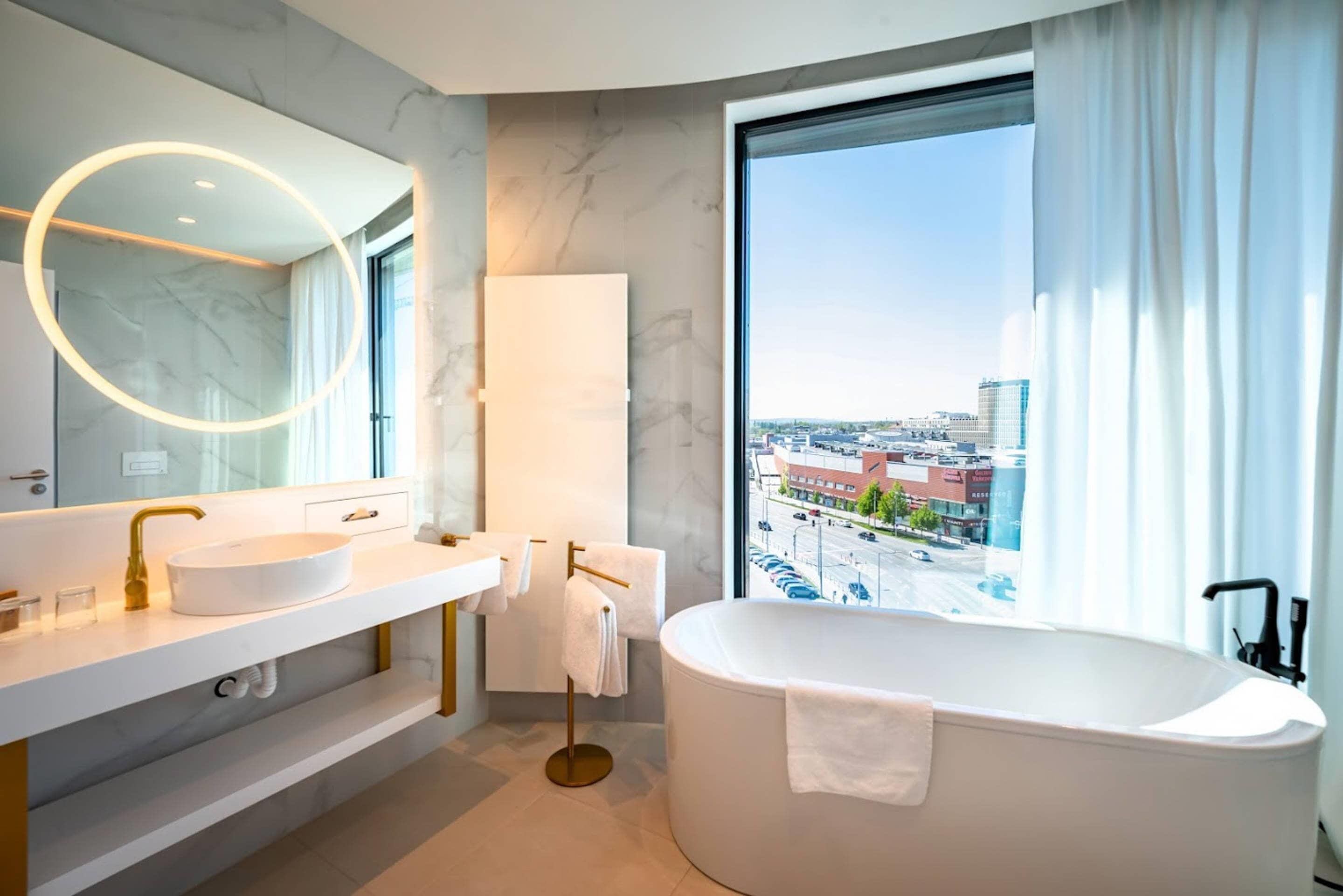 Un baño moderno con una bañera independiente, dos lavamanos, un espejo grande con luz circular y vistas a la ciudad a través de un ventanal.