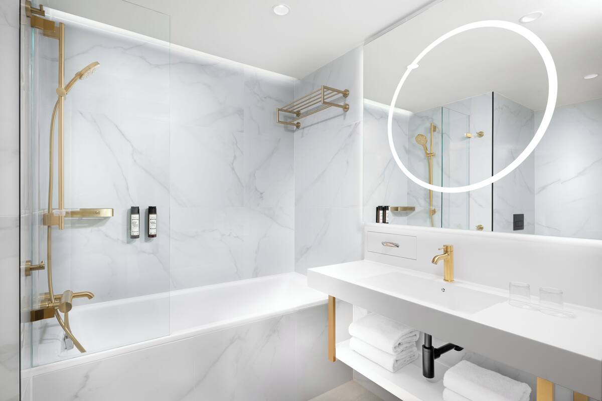 Une salle de bain moderne avec une douche à parois de verre, un mur en marbre blanc, un lavabo blanc avec des accessoires dorés et un grand miroir.