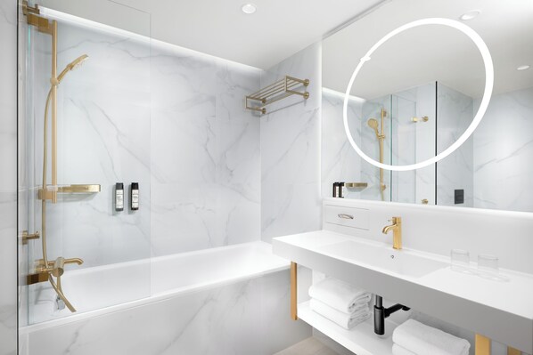 Un baño moderno con ducha envuelta en vidrio, pared de mármol blanco, lavamanos blanco con detalles dorados y un espejo grande.