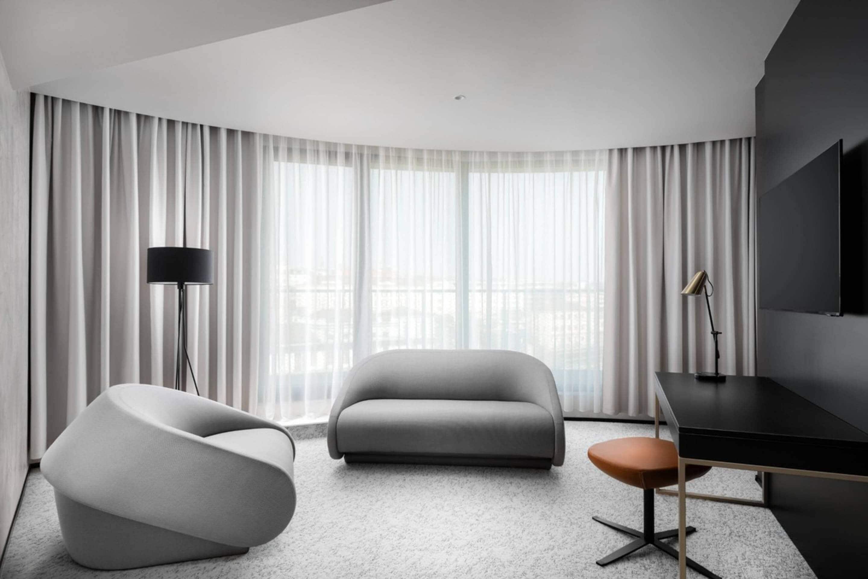A modern hotel room with a curved ceiling, a grey sofa, a white armchair, a desk with a chair, and a large window with sheer curtains.