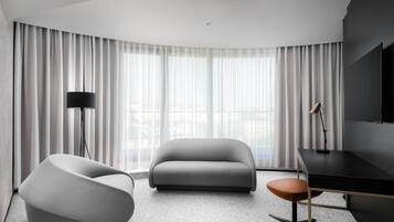 A modern hotel room with a curved ceiling, a grey sofa, a white armchair, a desk with a chair, and a large window with sheer curtains.