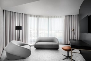 A modern hotel room with a curved ceiling, a grey sofa, a white armchair, a desk with a chair, and a large window with sheer curtains. - Clarion Congress Hotel Brno (Brno)