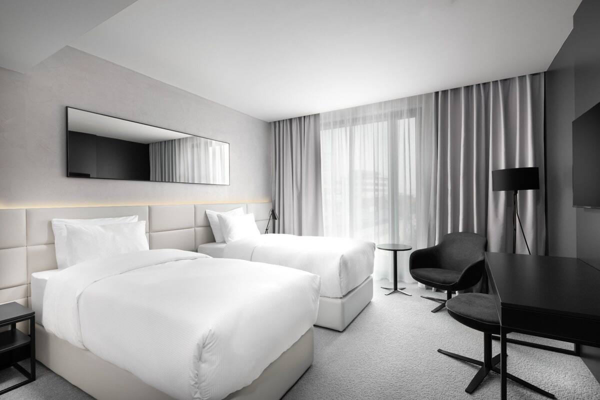 A hotel room with two beds, a desk, a chair, a TV, and a large window with curtains.