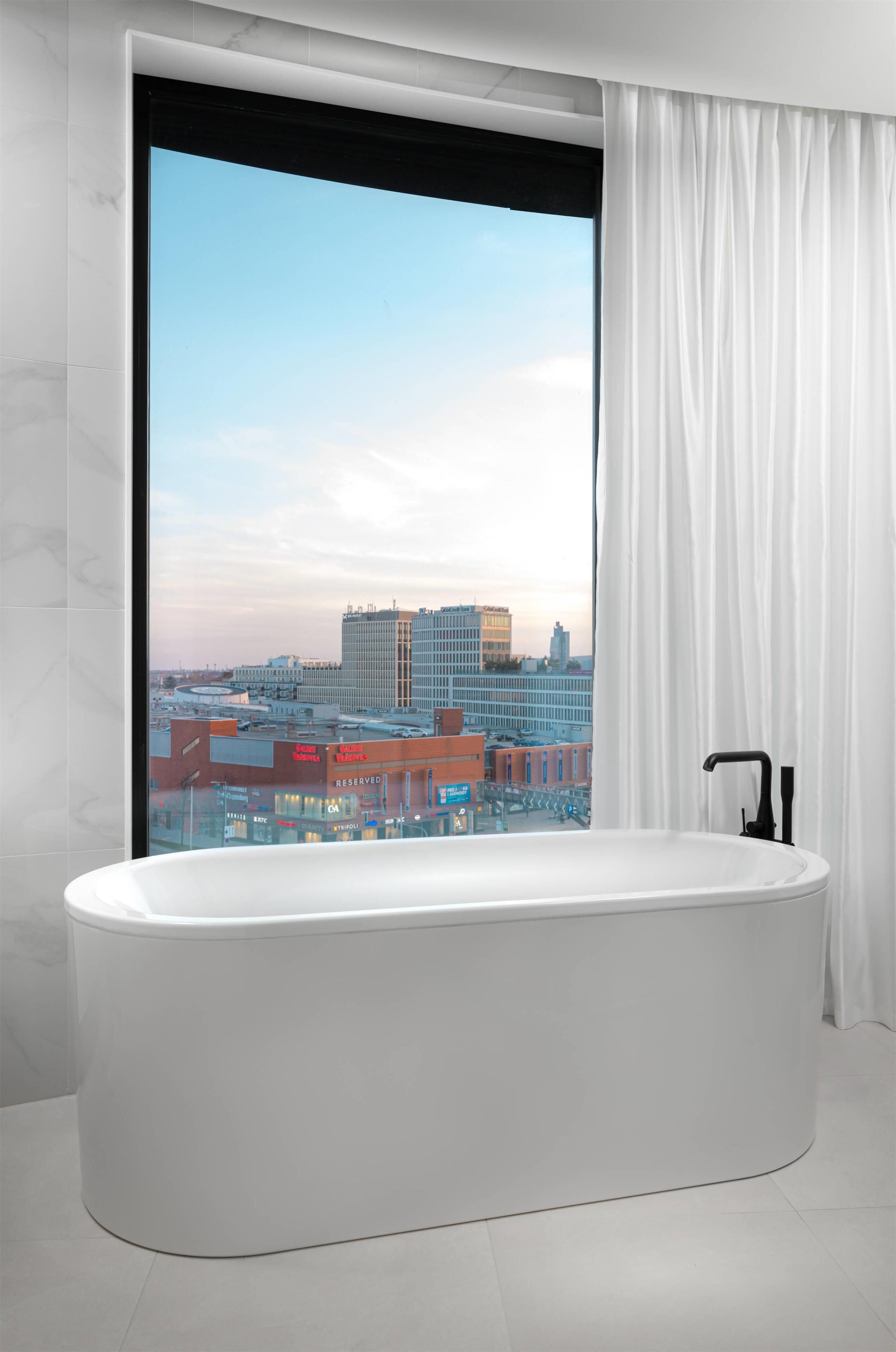 Un baño moderno con una bañera independiente, un ventanal con cortinas y vistas a un paisaje urbano.