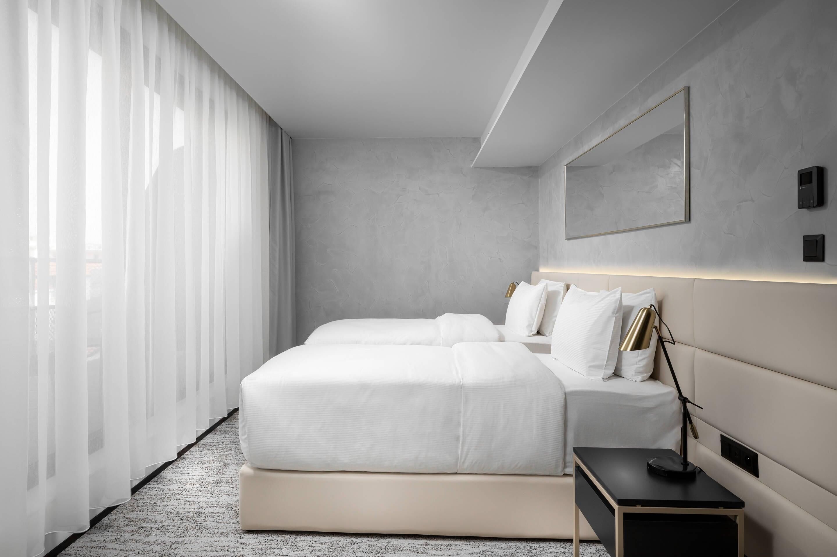 A modern hotel room with a large bed, bedside tables, and a large window with sheer curtains.