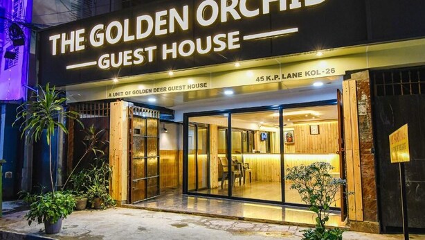 Exterior de The Golden Orchid Guest House, con una entrada moderna de puertas de vidrio, un área de recepción visible a través del vidrio y plantas en macetas en la entrada.