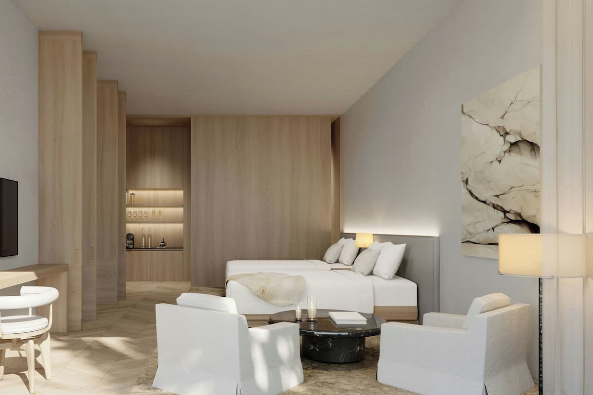 A modern hotel room with a large bed, two white armchairs, a coffee table, a TV, and a minibar.