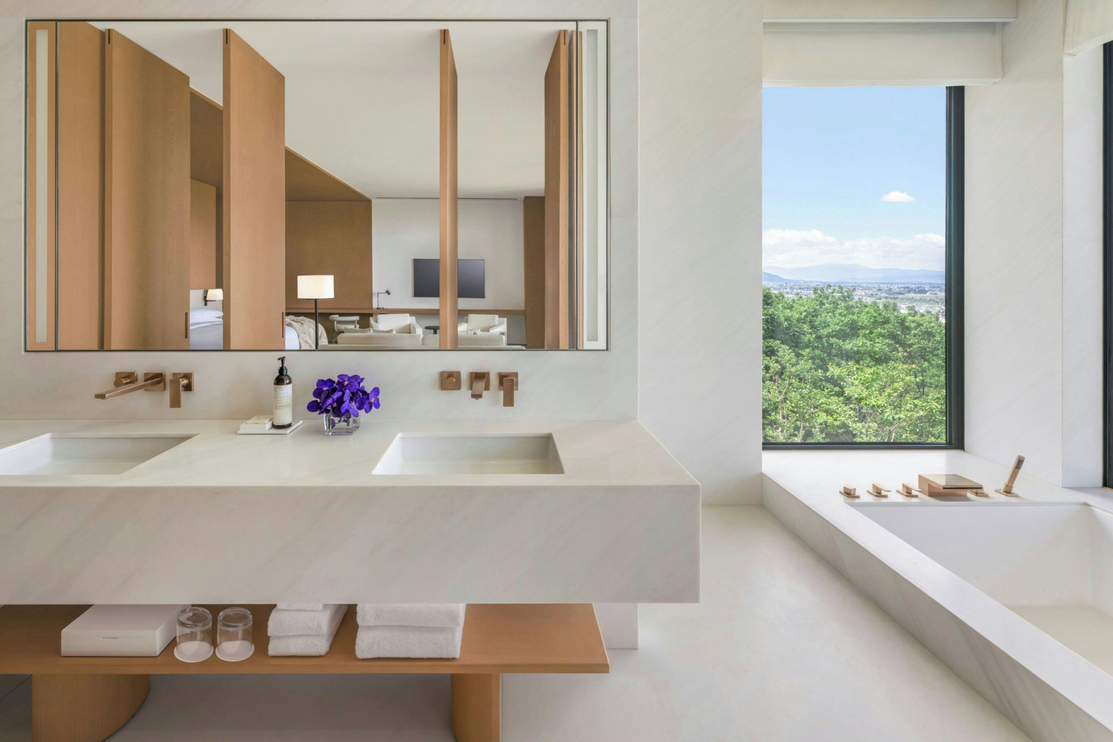 A modern bathroom with two sinks, a large mirror, and a bathtub with a view of greenery.
