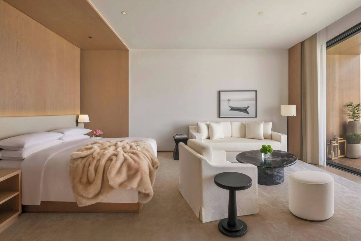 A modern hotel room with a bed, a sofa, a round coffee table, and a large window.