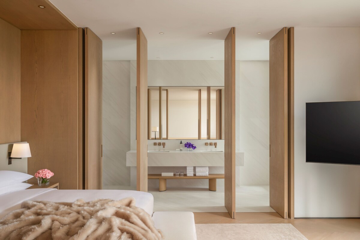 A modern hotel room with a large bed, a bedside table with a lamp, a TV, and a bathroom with a mirror, sink, and towel rack.