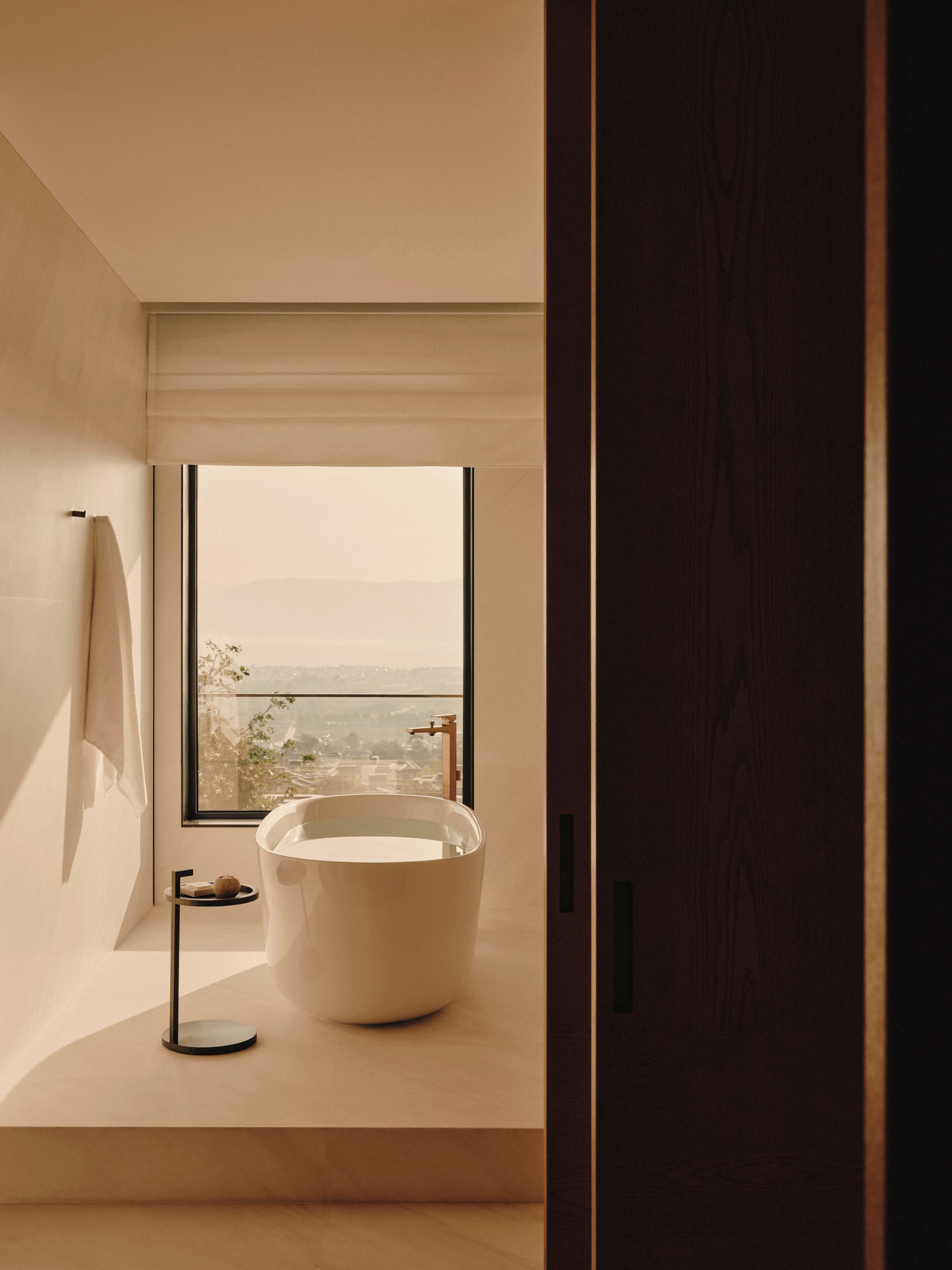 A modern bathroom with a large bathtub, a window with a view, and a wooden door.