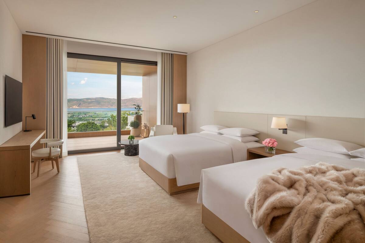 A hotel room with two beds, a desk, a chair, a TV, and a large window with a view.
