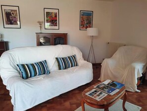 Un salon avec un canapé blanc, des coussins à rayures, une petite table en bois sur laquelle sont posés des magazines, un lampadaire et des œuvres d’art encadrées accrochées au mur.
