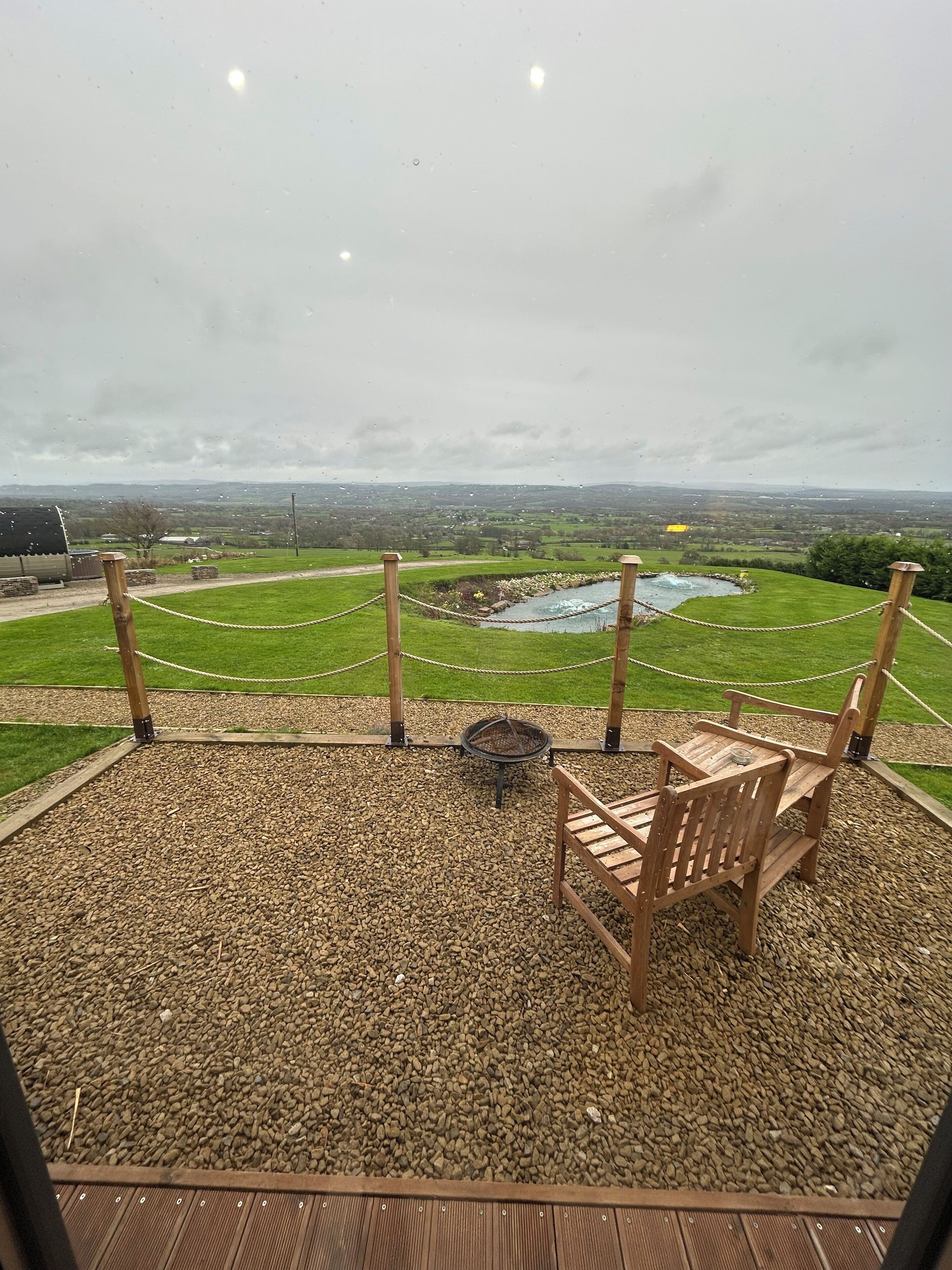 A covered outdoor seating area with a fire pit, wooden benches, and a view of a rural landscape.