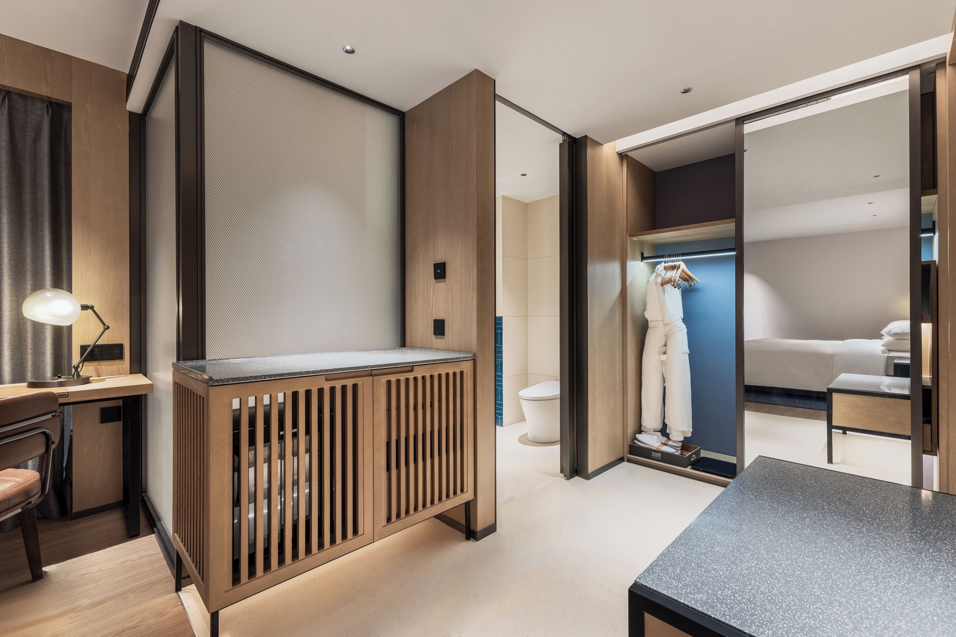 A modern hotel room with a wooden vanity, a desk with a lamp, a wardrobe, and a bed.
