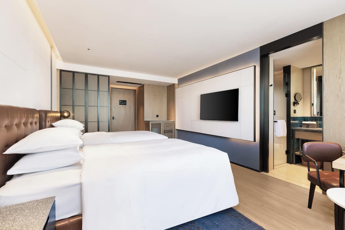 A hotel room with a large bed, a TV mounted on the wall, a seating area with a chair, and a bathroom visible through an open door.