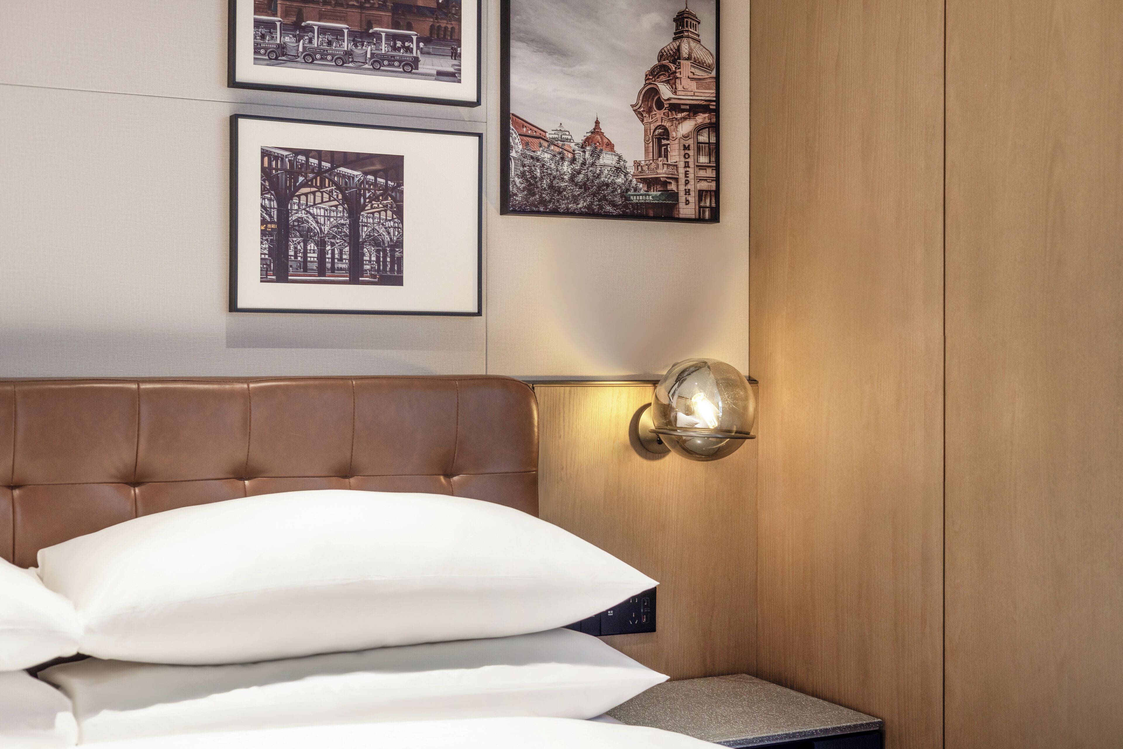 A hotel room with a brown headboard, a bed with white pillows, a small bedside table, and framed pictures on the wall.