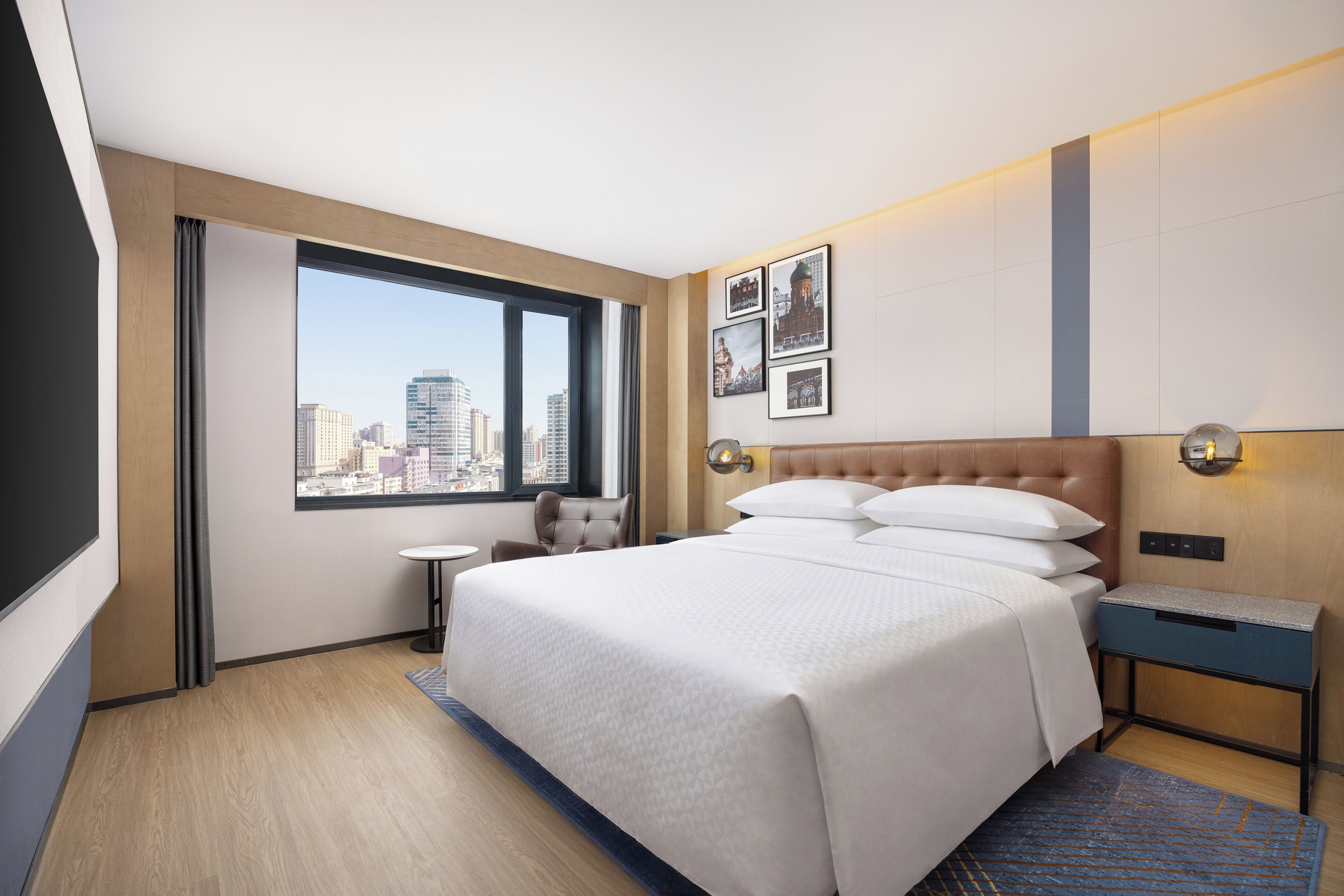 A hotel room with a large bed, a small table, a chair, a nightstand, and a city view through a large window.